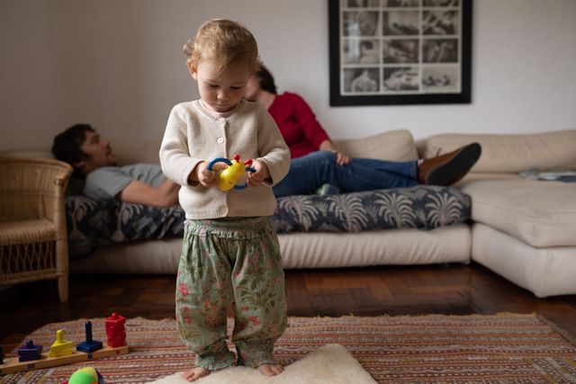 Front view of a Caucasian baby, standing on the carpet, playing with a toy, in the background both parents laying down on the sofa in a sitting room.