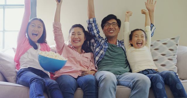 An Asian family celebrates a joyful occasion on the couch with popcorn and raised arms. - Download Free Stock Photos Pikwizard.com