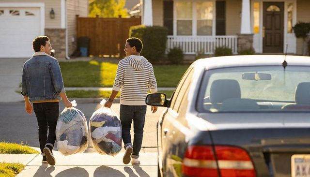 Young men carrying large clear bags of donated clothes down suburban sidewalk at sunset