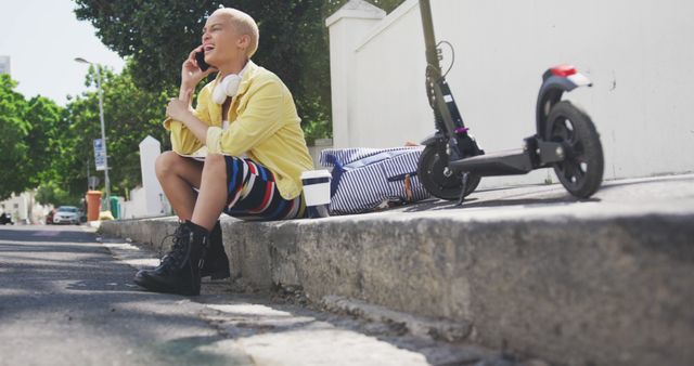 Young woman sits on curb with electric scooter, dressed casually, talking on a smartphone. This is perfect for city lifestyle themes, outdoor activities, technology use, and modern urban living contexts.