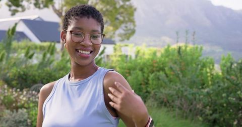 Smiling young woman adjusting hair in sunlit garden wearing glasses and casual tank top