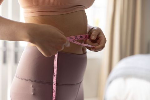 Woman measuring waist with pink tape wearing mauve activewear in soft light