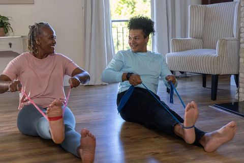 Diverse duo exercising with resistance bands on hardwood floor in sunlit living room