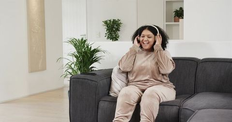 Curly Woman Enjoying Music on Headphones While Relaxing on Sofa in Cozy Neutral Living Room
