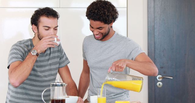 Two young men enjoying breakfast together in a modern kitchen. One man is drinking coffee while the other is pouring orange juice. Both individuals are dressed casually. The scene reflects a relaxed and cheerful atmosphere, making it perfect for promoting friendship, casual dining, morning routines, or lifestyle articles.