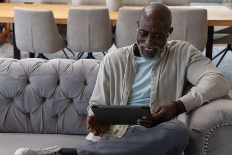 Senior african american man relaxing on tufted sofa using tablet in minimalist living room