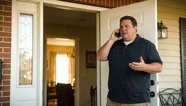 Casual homeowner talking on phone at front door, gesturing on sunny suburban porch