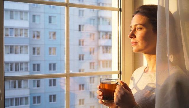 Woman holding tea gazing through sunlit apartment window at urban cityscape, morning calm