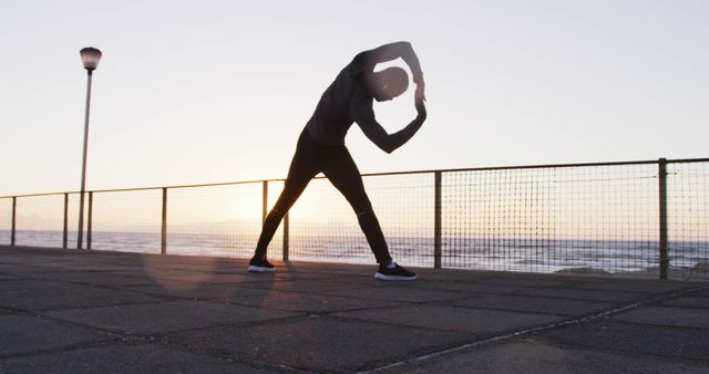 Image depicts a man stretching on an oceanfront boardwalk during sunrise. The man is extending his arms overhead, emphasizing flexibility and fitness. This image can be used for promoting health and wellness, fitness programs, outdoor exercise, and motivational content related to morning routines and staying active.