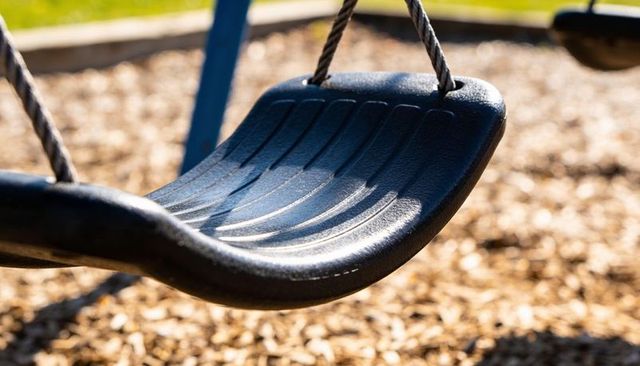 Sunlit black swing seat hanging on braided ropes over wood chip playground mulch
