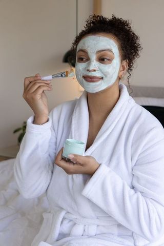 Curly-haired african american woman applying green facial mask in white robe for self-care
