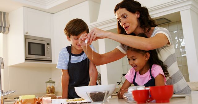 Mother cracking egg while baking with son and daughter in bright home kitchen. Children helping and learning to cook, smiling, having fun. Great for family-related content, learning activities, cooking blogs, kitchen appliance ads, nutritional articles.