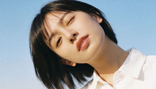 Winking young woman tilting face toward sun with serene expression and clear blue sky
