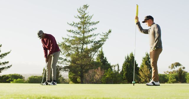 Two men are playing golf on a sunny day. One of them is focused on putting the ball while the other is holding a flagstick and watching attentively. Ideal for use in content related to golfing, outdoor sports, leisure activities, and promoting healthy lifestyle.
