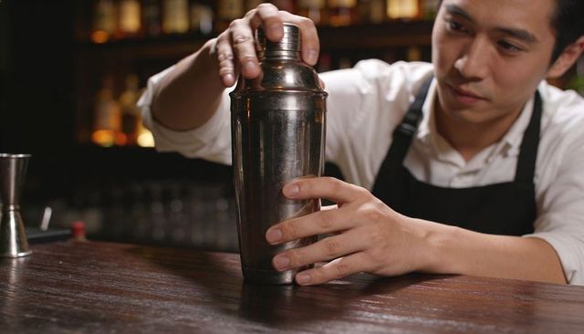 Shaking cocktail shaker behind wood bar counter with jigger and warm backlight
