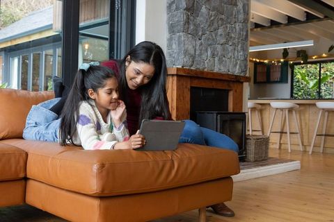 Indian mother and daughter using tablet on tan leather sofa by fireplace in openplan home
