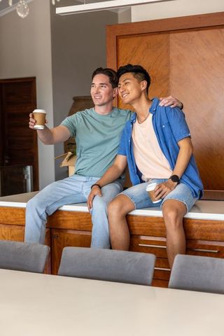 Diverse male friends sitting on kitchen counter holding coffee cups while unpacking new home