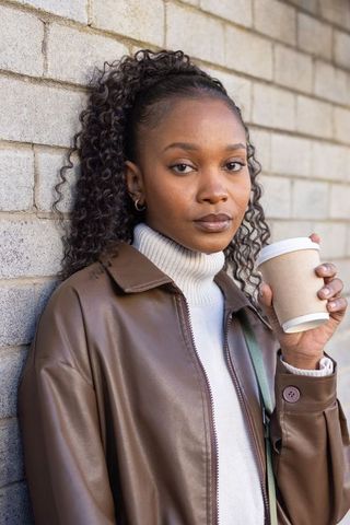 African woman leaning on brick wall holding coffee cup in leather jacket streetstyle