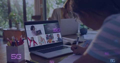 Child attending virtual classroom on laptop while writing homework at home workspace