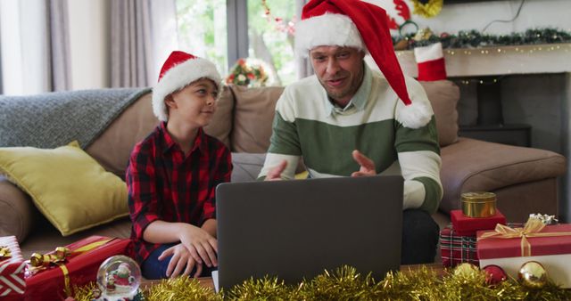 Father and son sitting in a decorated living room, wearing Santa hats and having a video call on a laptop. Perfect for holiday greetings, family connection, and Christmas celebration themes. Ideal for promoting family traditions, virtual meetings, and festive holiday products.