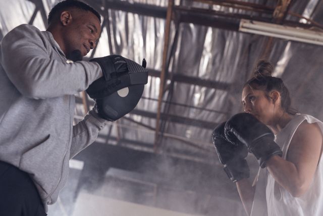 Low angle view of a dedicated multiracial male and female boxers practicing in a gym. Both are wearing boxing gloves and appear focused on their training. This image can be used for promoting fitness programs, boxing classes, sportswear, or motivational content related to health and fitness.