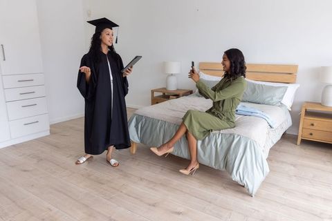Diverse mother recording graduate daughter delivering commencement speech in bedroom