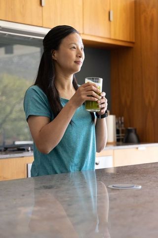 Mature asian woman holding green smoothie in sunlit modern kitchen, wellness lifestyle