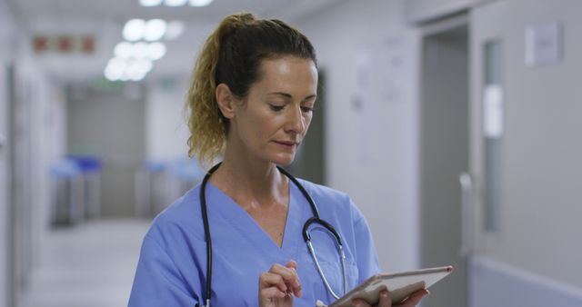 Female nurse wearing blue scrubs standing in hospital corridor while using a digital tablet. Ideal for illustrating modern healthcare, medical professionals utilizing technology, patient management, and hospital environments.