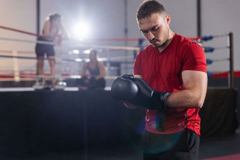 Male boxer adjusting black glove near boxing ring while trainers watching in backlit gym