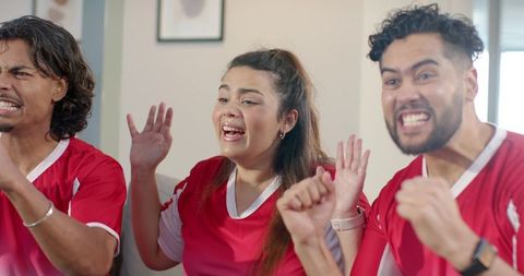 Diverse friends cheering and leaning forward on sofa wearing red soccer jerseys