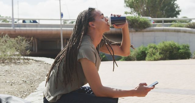 Fashionable biracial man with dreadlocks using smartphone and drinking coffee. Street style, modern urban lifestyle and communication.