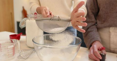 Two women sifting flour into glass mixing bowl while baking together in bright kitchen