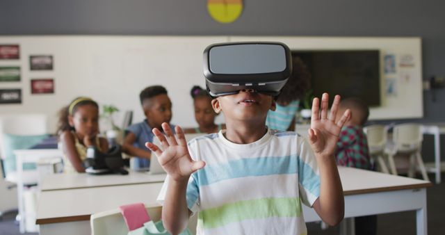 African American boy experiencing virtual reality with a VR headset in a classroom. Other children in background engaging with technology. Illustrates the integration of modern technology into education, making learning more interactive and engaging. Ideal for articles on educational technology, future of learning, or promotional materials for innovative teaching methods.