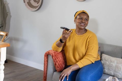 African american woman smiling while sending voice message from sofa in cozy living room