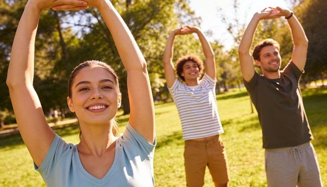 Sunlit park group stretching, smiling young adults raising arms during outdoor workout