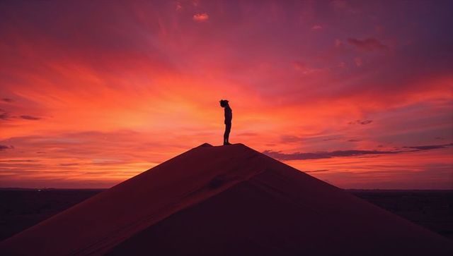 Standing silhouetted hiker on dune crest at dramatic crimson sunset conveying solitude