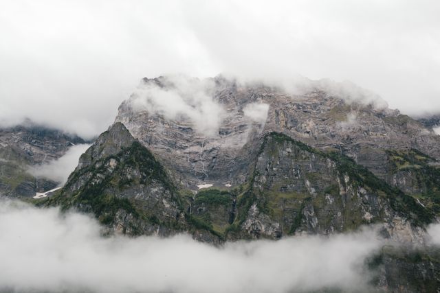 A scenic view of a mountainous range partially covered in mist and clouds. The peaks display rugged gray rocks while lush green foliage fills the lower sections. Ideal for use in travel brochures, nature magazines, wall art, and websites promoting outdoor adventures and serene natural landscapes.