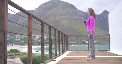 Mature woman exercising with dumbbells on coastal balcony overlooking beach and mountains