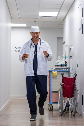 Joyful doctor walking hospital corridor holding coffee and celebrating with raised fist