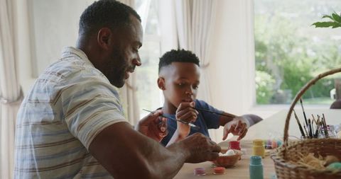 African american dad and son decorating easter eggs at sunlit kitchen table