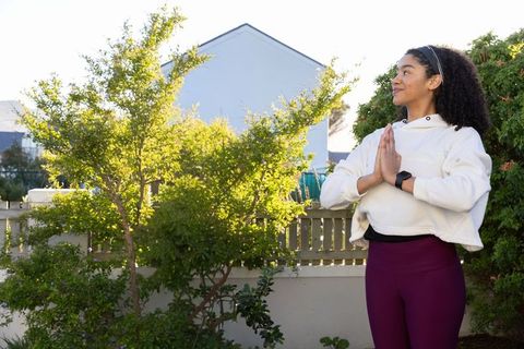 Young woman practicing mindfulness yoga in sunny backyard wearing hoodie and smartwatch