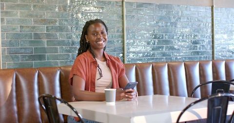 African woman smiling while holding smartphone in modern cafe booth, casual rust top