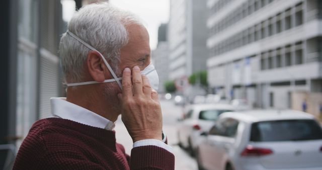 Elderly man with white hair seen adjusting protective mask while standing in an urban environment. Everyone can use this for concepts related to health, safety during pandemics, or everyday urban life in older adults.
