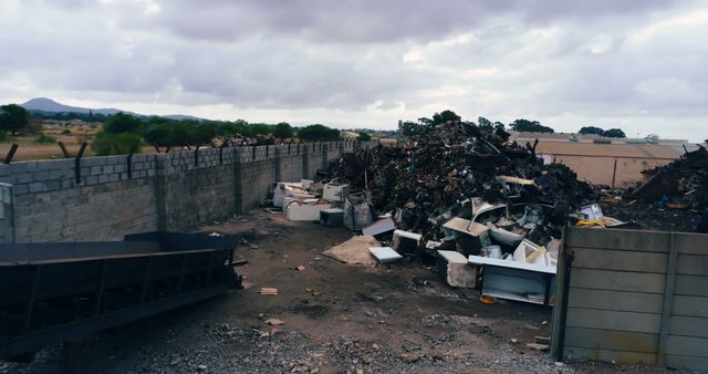 Large pile of industrial waste piled against a concrete wall under a cloudy sky. The scene includes discarded appliances and scrap materials, highlighting environmental pollution. Useful for topics related to waste management, environmental issues, industrial decline, and environmental awareness campaigns.