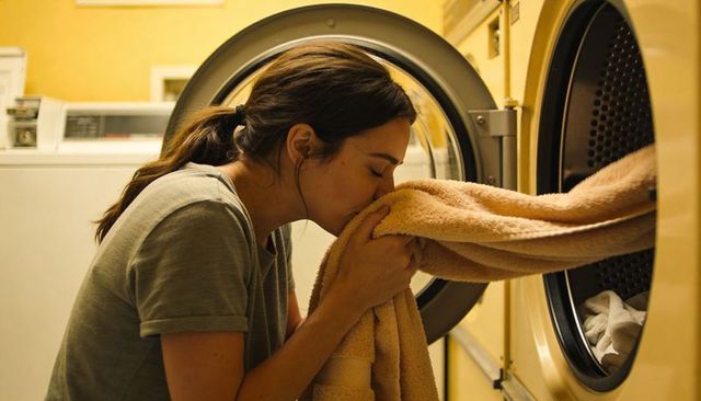 Woman smelling warm towel emerging from dryer in cozy laundromat routine, savoring fresh linen