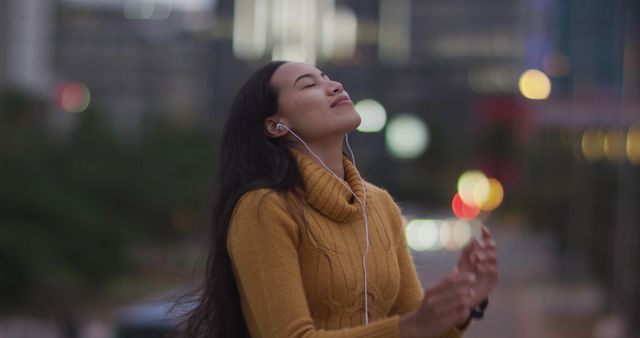 Young woman wearing yellow sweater enjoying music with her eyes closed in the evening cityscape. Perfect for articles on leisure, relaxation, urban lifestyle, music enjoyment, and stress relief. Enhances content related to personal well-being, night life, and serene moments in bustling cities.