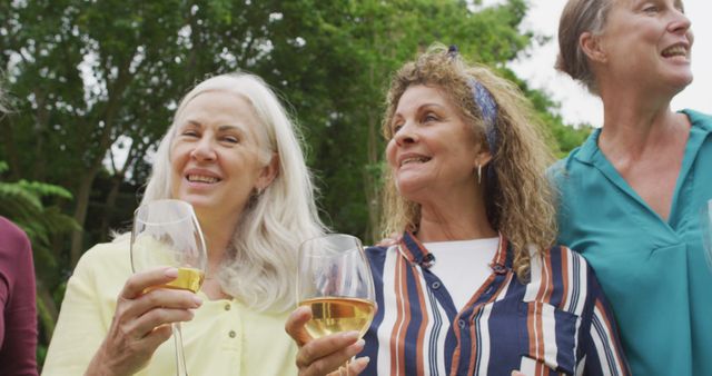 Three women enjoying a drink together outside, smiling and engaged in conversation. All are holding wine glasses and appear happy, suggesting they are at a social gathering or celebration. Ideal for themes related to social events, friendship, celebrations, senior activities, and outdoor gatherings.