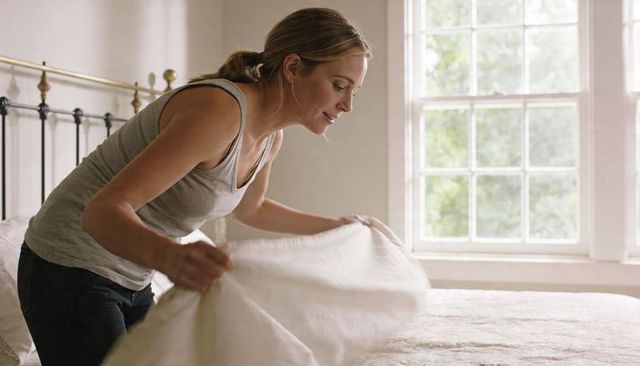 Woman making bed in bright minimalist bedroom with brass headboard and sunlit window