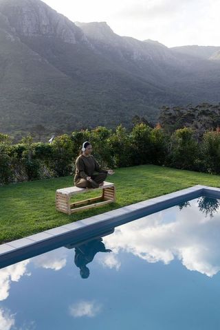 Mid-adult asian woman meditating poolside wearing headphones with mountain reflection