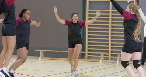 Diverse female teammates celebrating and jumping on indoor gym court in team uniforms